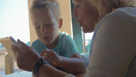 En-La-Ciudad-De-Perea,-Grecia,-Se-Sienta-Una-Abuela-Con-Su-Nieto-Y-Le-Enseña-Cómo-Usar-La-Tableta.