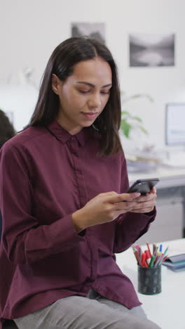 video vertical de una mujer biracial sonriente usando un teléfono inteligente en el trabajo