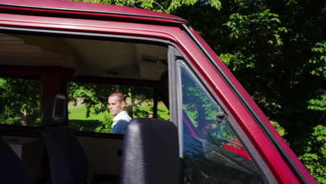 Young-man-gets-in-beatiful-red-retro-bus,-van-and-putting-on-seat-belt-on-an-sunny-summer-day