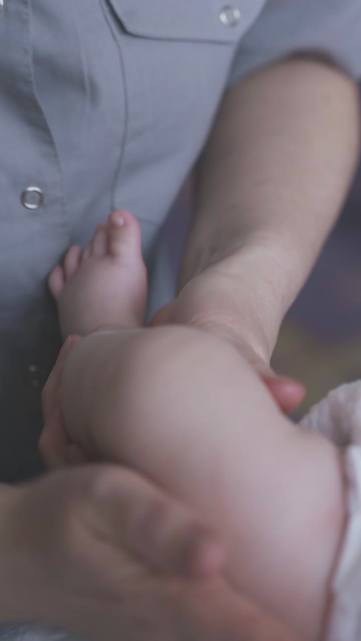 small infant boy in white diaper lies on baby changing table and skilled nurse performs legs massage close view