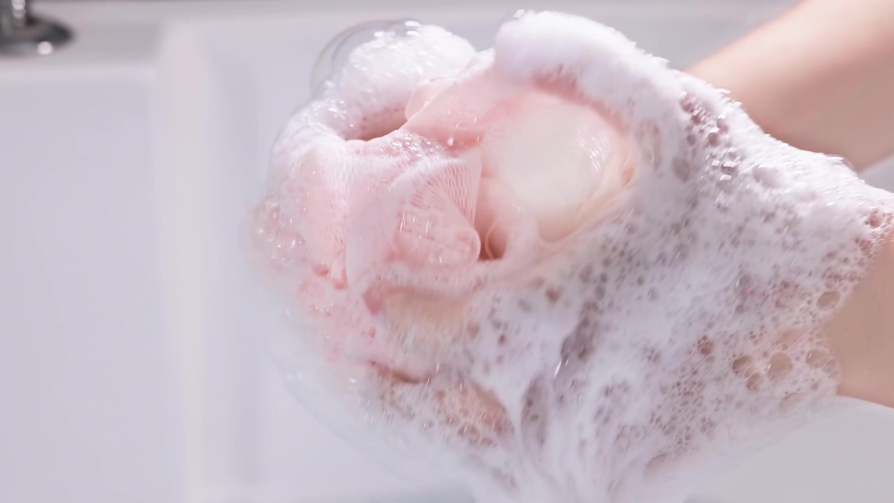 Primo piano delle mani di una giovane donna che versa shampoo o bagnoschiuma su una spugna a rete da bagno, per la pulizia, la cura del corpo, l'igiene personale e la salute, usando un prodotto di sapone liquido per la pulizia del corpo nella vasca da bagno.