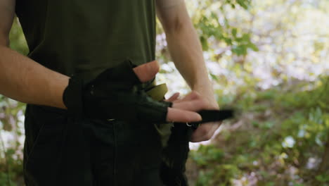 close up view of young man wearing military glove, clapping strap under dappled sunlight filtering through foliage canopy in dense forest clearing, veins visible on wrist, blurred trunks beyond