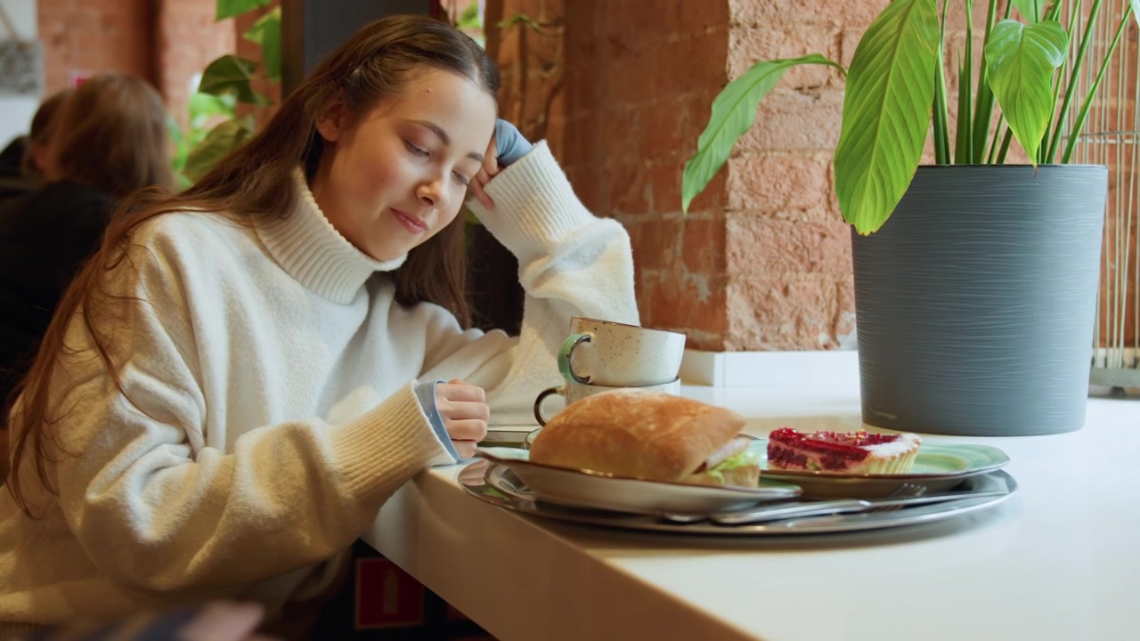 Close up of young woman resting head on hand at table with food and drink. Woman in white sweater appears relaxed surrounded by plate sandwich cup and dessert. Warm indoor setting with brick