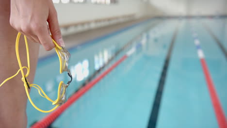 holding yellow swimming goggles, person standing by indoor swimming pool, copy space