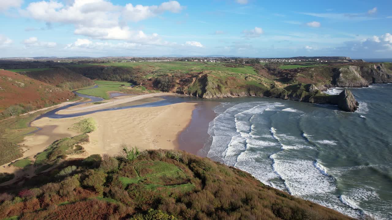 Drone gliding above a sandy beach, rugged cliffs with waves below and grassy headlands beyond at Three Cliffs Bay on the Gower Peninsula in Wales