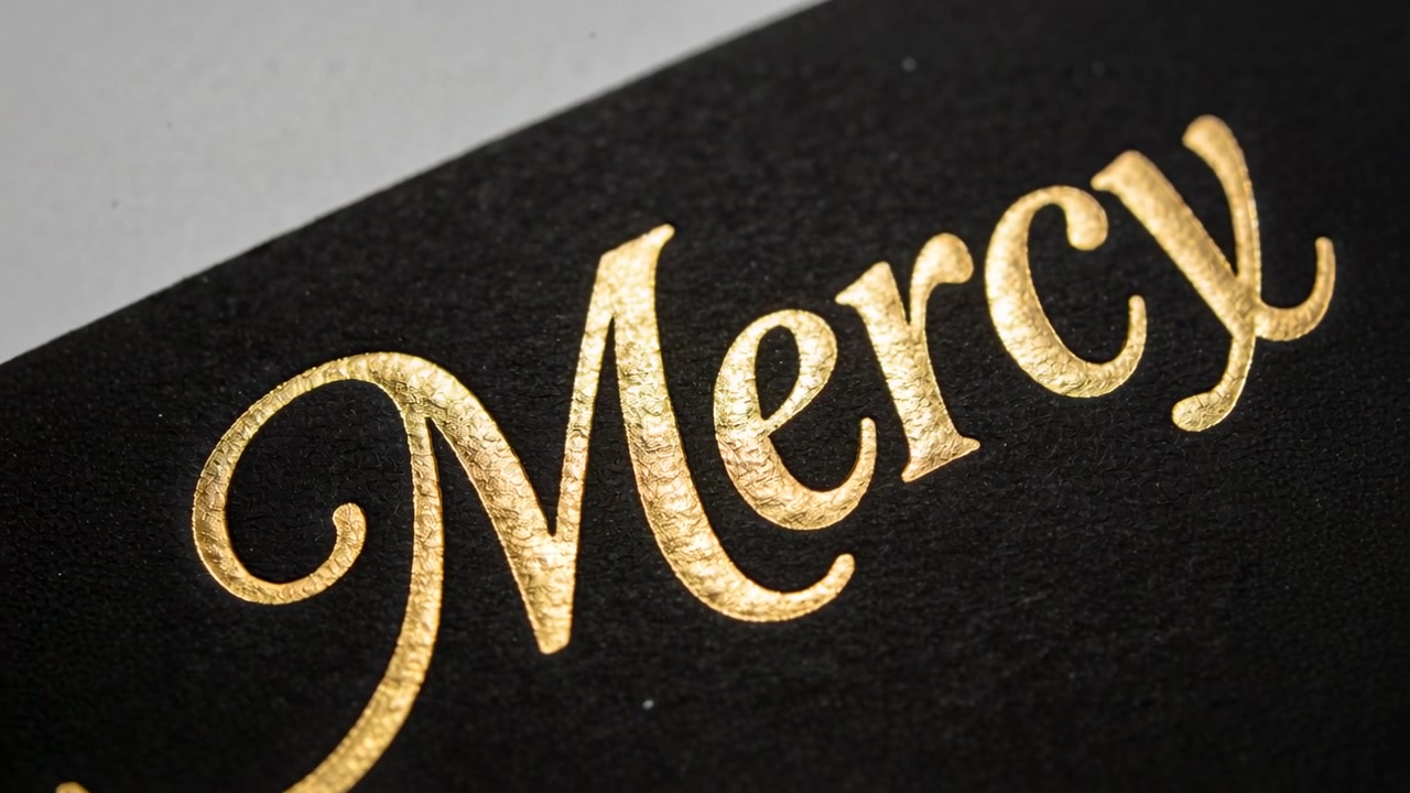 Inclinando la fotocamera, rivelando la parola "Mercy" con lamina d'oro su carta nera testurizzata, evidenziando la goffratura metallica.