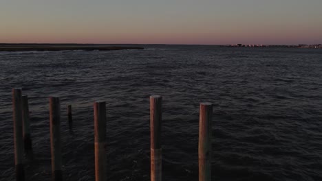 Drone-flies-above-wooden-support-poles-in-open-ocean-at-dusk,-Chincoteague-Island-Virginia,-slow-motion