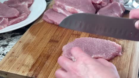 Cutting-off-white-fat-from-raw-slice-of-pork-with-chef's-knife---solid-fat-being-sliced-off-from-slab-of-meat-in-kitchen-on-wooden-cutting-board---chopping-off-fat-at-home-inside---schnitzel-recipe