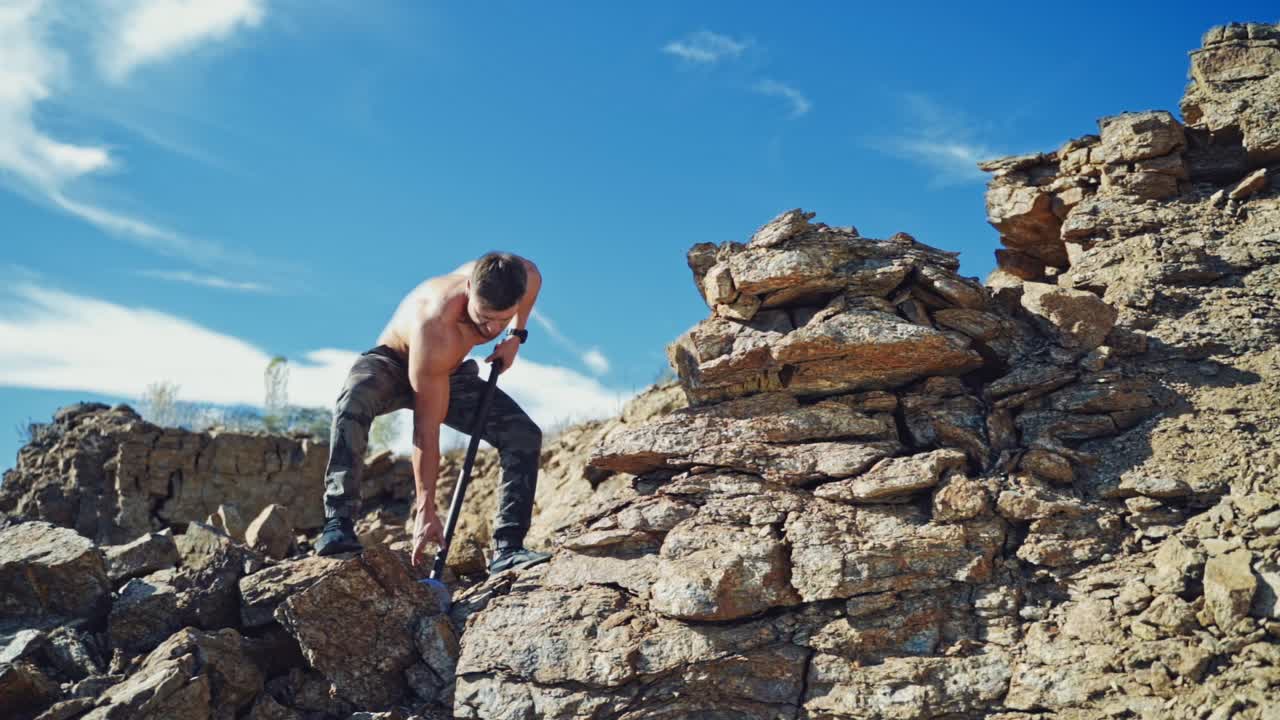 Athletic man exercises with a big hammer in canyon. Muscular man standing on rock and breaking stones with a sledgehammer under the blue sky. Slow motion.