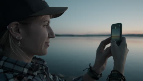 happy woman photographing sunset over lake with phone