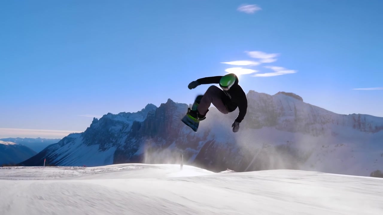 Uno snowboarder si muove su un luminoso pendio montano coperto di neve sotto un cielo azzurro limpido, con cime distanti, forte luce solare, lunghe ombre e un ampio panorama scenografico.