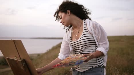 Beautiful-curly-woman-artist-concentrated-on-painting-process.-Working-on-future-picture,-using-a-paintbrush-applies-oil-paints.-Outdoors