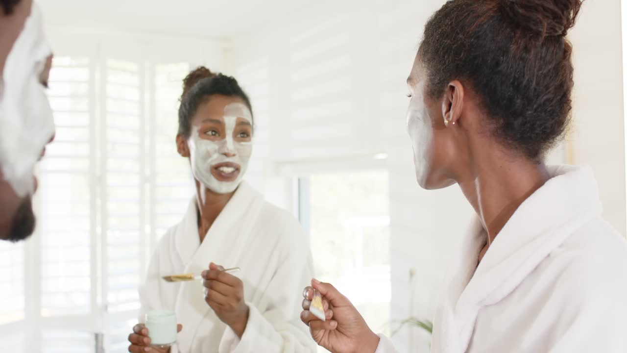 Afroamerikanische Frauen in weißen Roben am Schminktisch sehen in den Spiegel, tragen Masken mit Goldpinsel aus dem Tiegel auf.