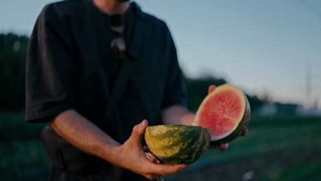 Nahaufnahme-Eines-Mannes-In-Einem-Schwarzen-T-Shirt,-Der-Auf-Einem-Feld-Auf-Einer-Farm-Eine-Kleine-Wassermelone-Aufschneidet-Und-Die-Hälfte-Seinem-Freund-Gibt