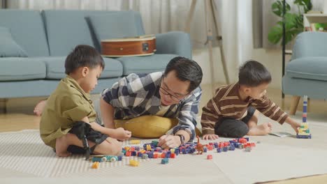 full body of asian father and sons playing the construction set colorful plastic toy brick at home. the kids assemble plastic building blocks sitting on a mat, a father lying on the floor talking to his sons