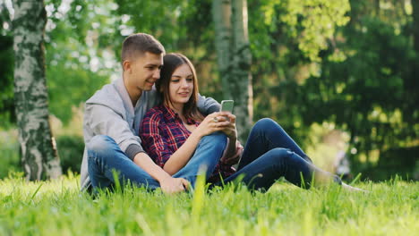 Young-Couple-Resting-Together-In-The-Park-Play-On-Your-Phone-Smiling-Guy-Hugs-Girl