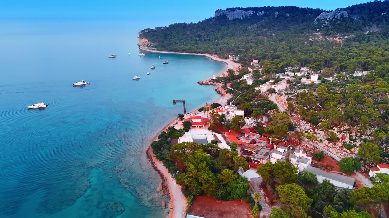 Footage over the coast of the Mediterranean Sea with houses and multiple trees. Some boats are on the waterscape near the shore. Kemer, Turkey