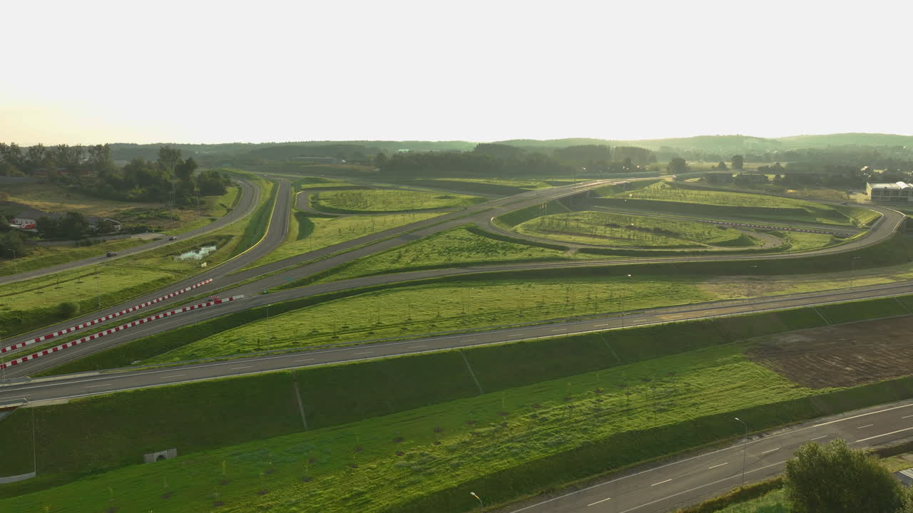 Expansive aerial view of a large, modern highway interchange featuring multiple ramps and sweeping curves. Golden sunlight bathes the green grassy slopes