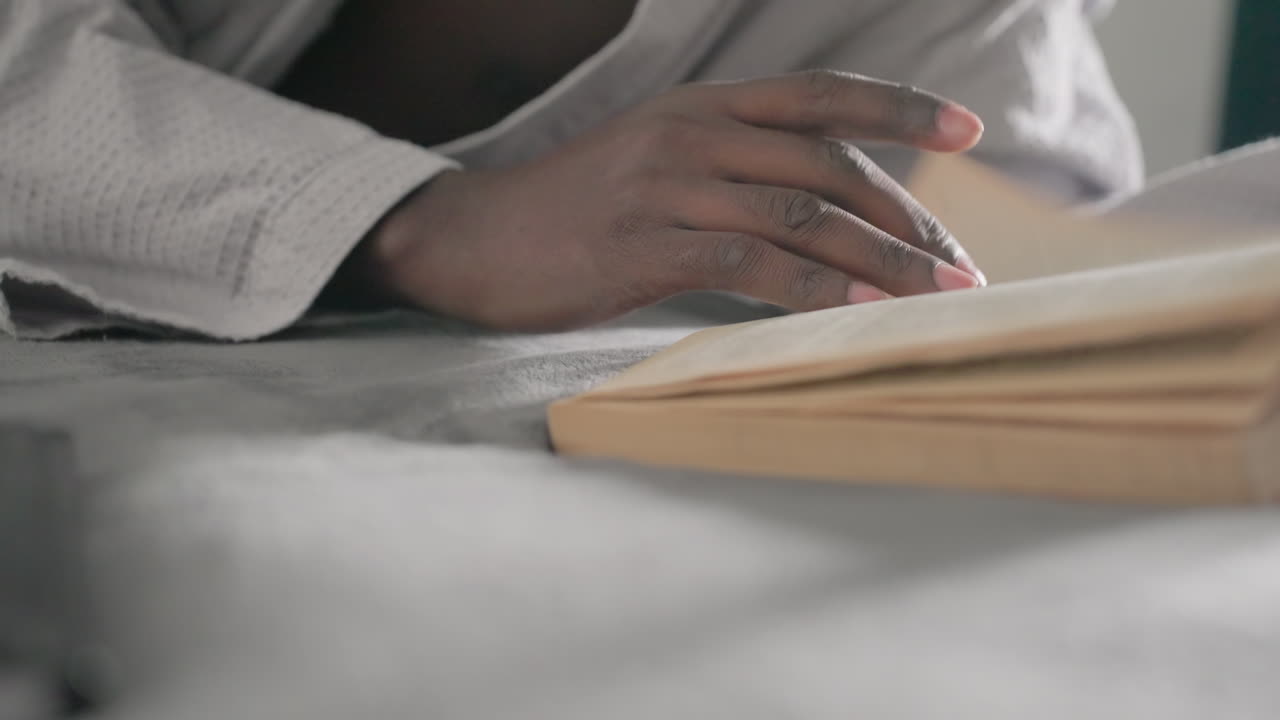 Close-up of hand turning pages of large book while lying on bed, focused on reading, soft light streaming through window, creating peaceful, introspective atmosphere in tranquil indoor setting