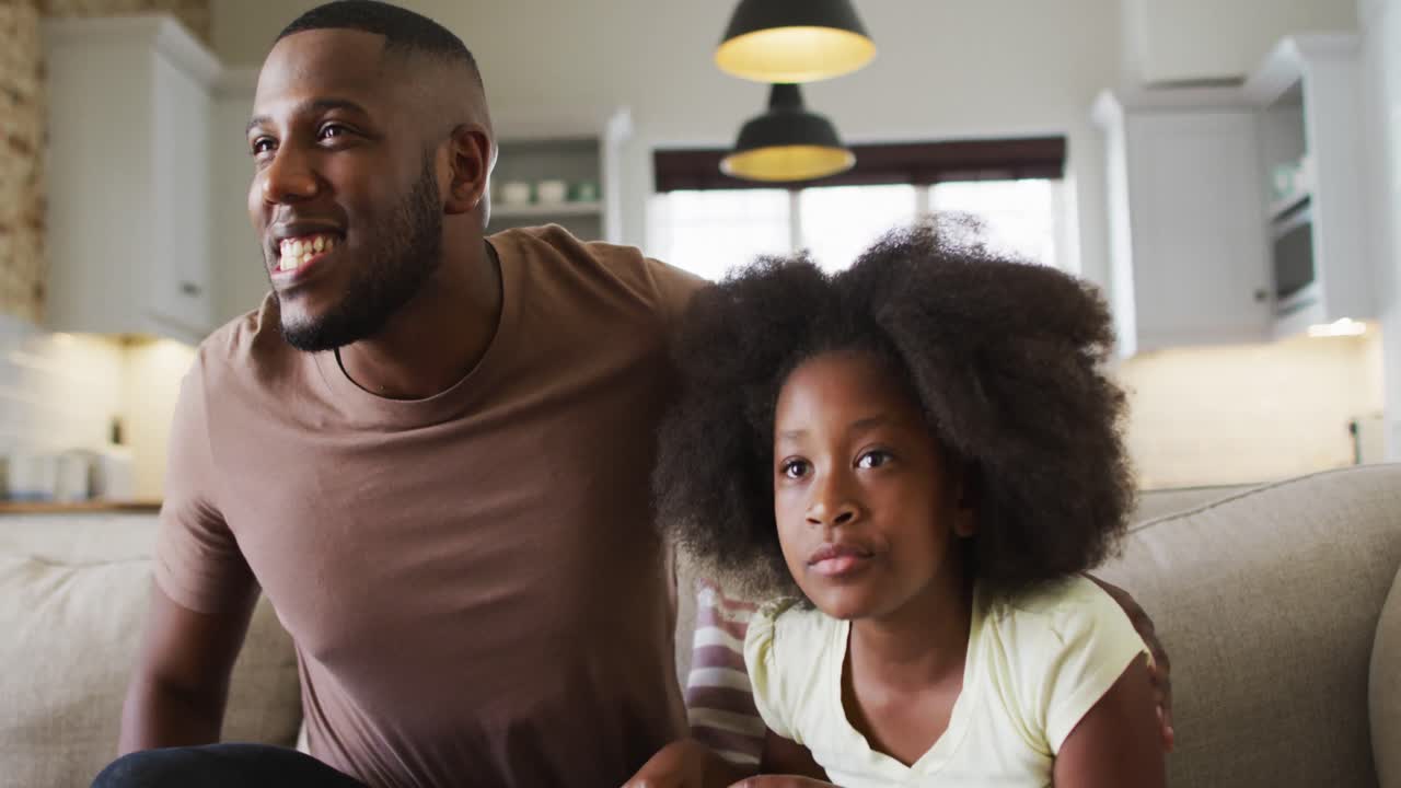 Figlia afroamericana e suo padre sul divano che guardano la TV, mangiano popcorn e fanno il tifo.