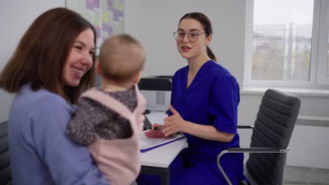 über-Die-Schulter-Ein-Selbstbewusstes-Brünettes-Mädchen-Mit-Ihrer-Kleinen-Tochter-Ein-Baby-Bei-Einem-Termin-Mit-Einem-Kinderarzt-In-Einer-Blauen-Uniform-Ein-Mädchen-Mit-Brille-Selbstbewusstes-Mädchen-Arzt-Kinderarzt-Kommuniziert-Mit-Einer-Jungen-Mutter-Und-Ihrer-Kleinen-Tochter-Baby-In-Einer-Modernen-Klinik