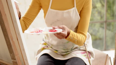 woman painting on canvas at home, wearing yellow sweater and apron, smiling