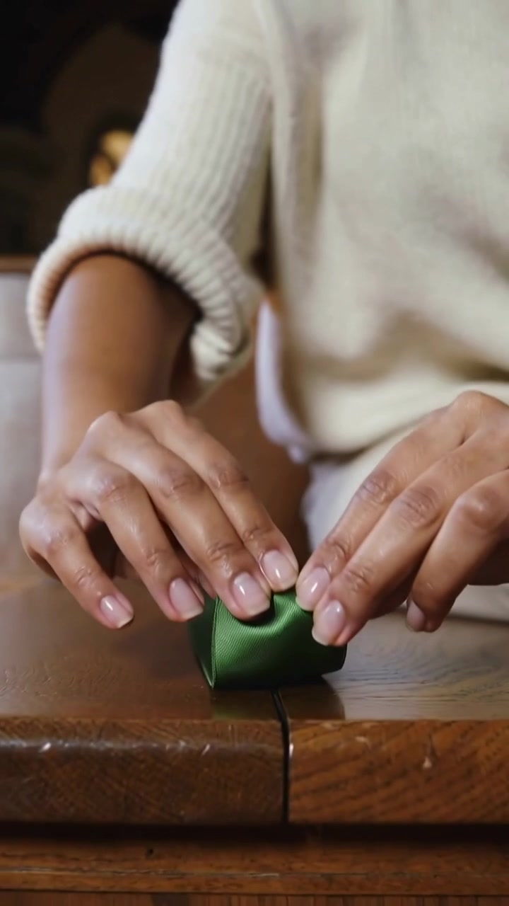 Video verticale: Nastro che si srotola, donna in top color crema a casa che piega un nastro verde su una cucitura per fare un fiocco