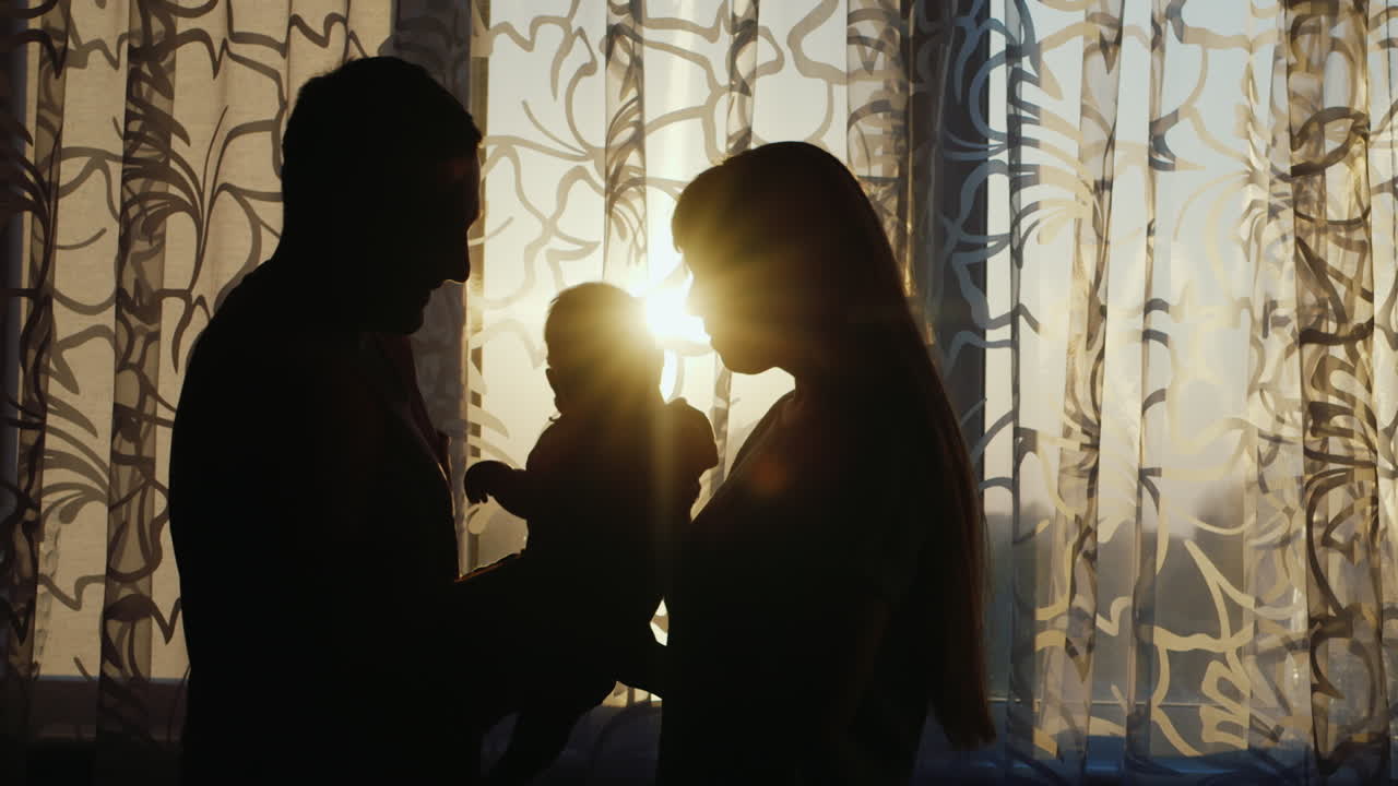 행복한 가족의 실루엣은 해질녘 창가에 있는 집으로 아이를 부드럽게 팔에 안고 있습니다