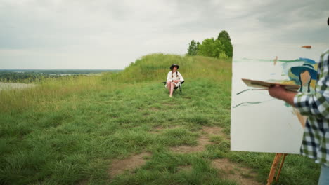 a painter in a hat and checkered shirt, holding a palette, is focused on creating a masterpiece in a grassy field. in the background, a woman in a hat and white dress sits , posing for the painting