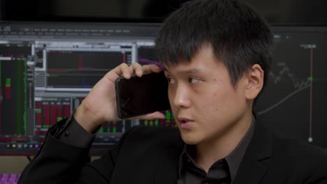 hombre chino preocupado hablando por teléfono con pantalla de mercado de valores en el fondo