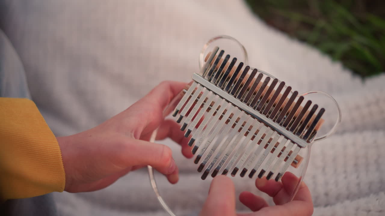 맑은 칼림바를 야외에서 연주하는 하얀 손, 부드러운 담요 위에 노란 소매, 투명한 음쇠를 뜯는 손가락, 아늑한 오후의 휴식과 마음 챙김 수련을 떠올리게 하는 부드러운 멜로디