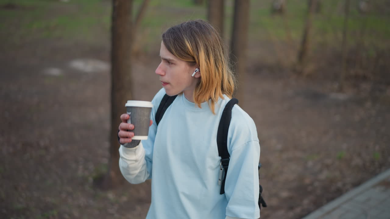 Estudiante joven de pie con un café en un parque tranquilo, con auriculares a la vista, mochila al hombro, expresión pensativa, sudadera de color pastel, luz de la mañana, árboles sin hojas, vaso desechable, cinematográfico
