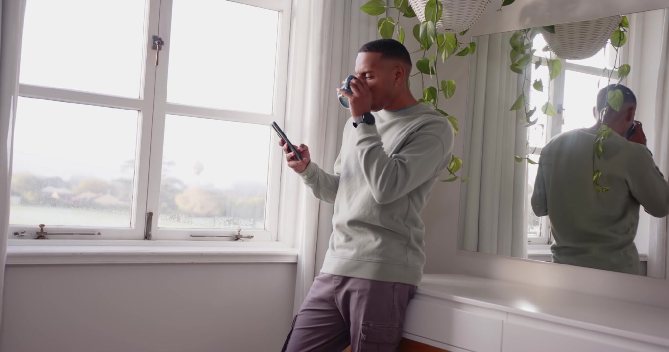 Uomo adulto africano appoggiato a una panca vicino a una finestra, che scorre il telefono dopo un avviso, sorseggiando da una tazza, con spazio per il testo.
