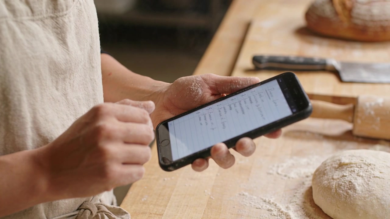 Tapping female baker in light apron consulting smartphone at wooden workbench, locating dough step
