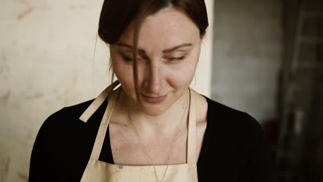 Portrait-of-young-good-looking-woman-wearing-apron-sitting-in-pottery-and-making-clay-vase-on-potter-wheel-using-hands.-Woman-smiling-to-a-camera