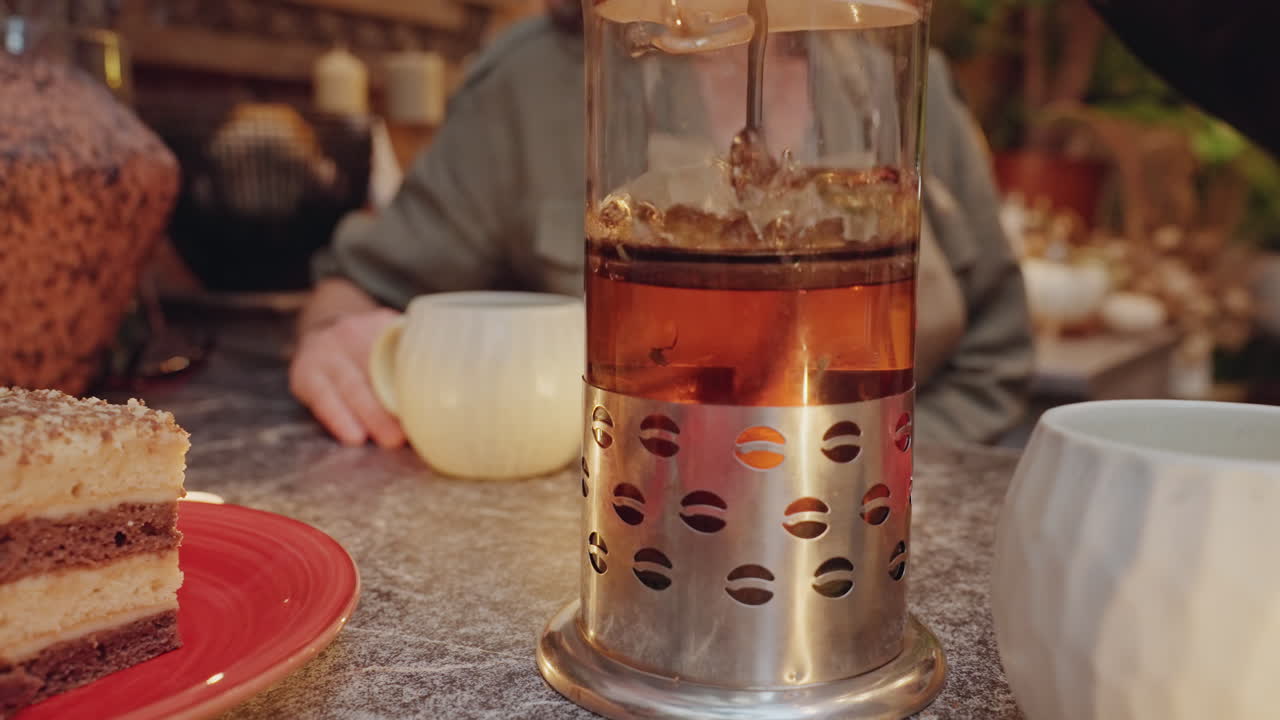 Floristin genießt Tee und Kuchen, Hände bedienen eine French Press, Keramiktasse neben einem geschichteten Kuchenstück auf rotem Teller, Kerzenschein, rustikale Werkstatt voller Pflanzen und Blumensträuße, gemütliche Abendpause.