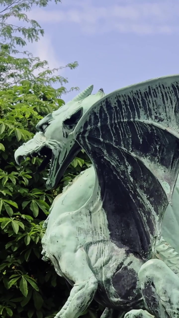 Primo piano verticale dell'iconica statua del drago verde sul Ponte dei Draghi di Lubiana, che rivela la sua patina metallica invecchiata, le grandi ali e la forma mitica sullo sfondo della verdeggiante vegetazione estiva