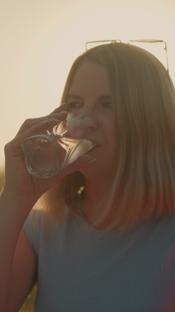 mujer de camiseta azul bebe un refrescante vaso de agua al aire libre, rodeada de verde sereno y bañada en la cálida luz del sol, disfruta de un momento de paz en la naturaleza, con un fondo escénico borroso