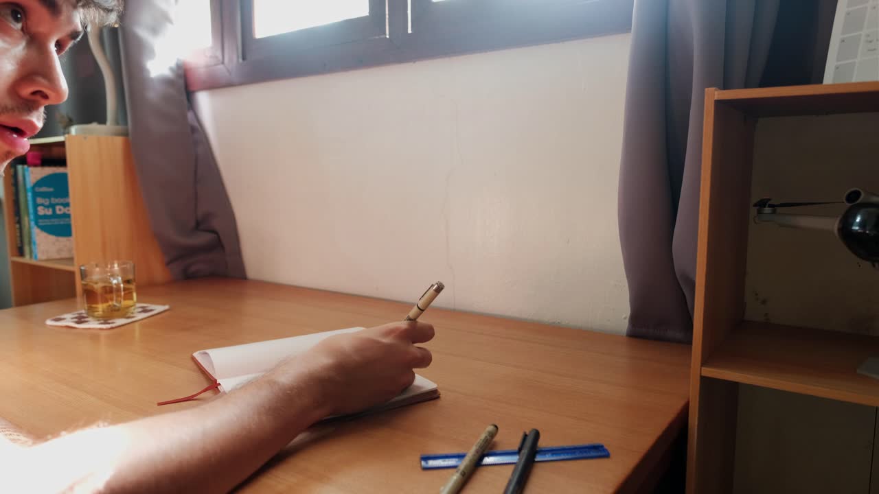 Side medium shot of male student sitting at a sunlit wooden desk, writing in open journal at calm bedroom workspace