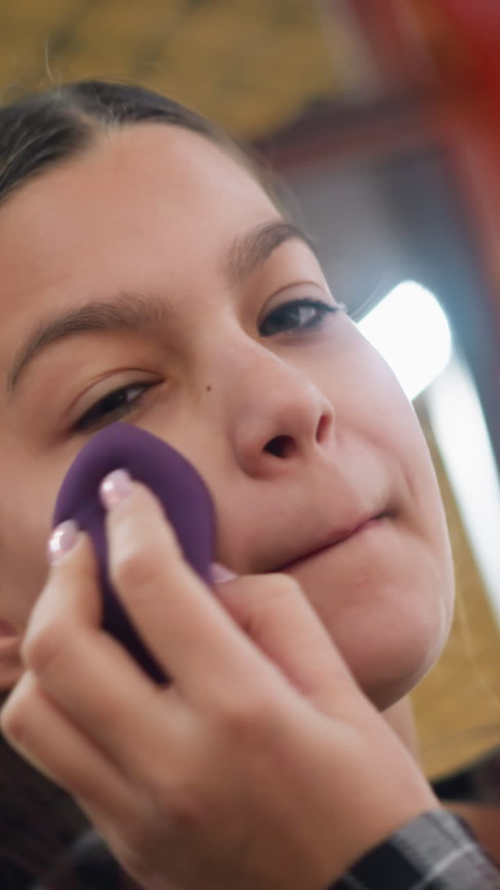 Primer plano de una adolescente aplicándose suavemente maquillaje con una esponja; sus manos presionan una esponja de belleza morada contra sus mejillas; luz natural, expresión concentrada, manicura visible, cosméticos sutiles, tranquilidad