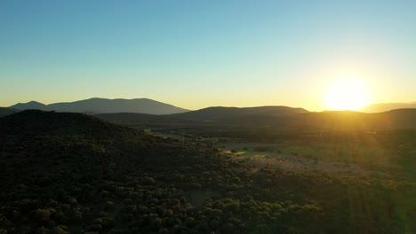 Backlit-drone-filming-in-the-spectacular-Tiétar-Valley-where-we-see-hills-full-of-green-trees,-meadows,-a-mountain-system-in-the-background-with-a-blue-sky-and-striking-flashes-produced-by-the-sun