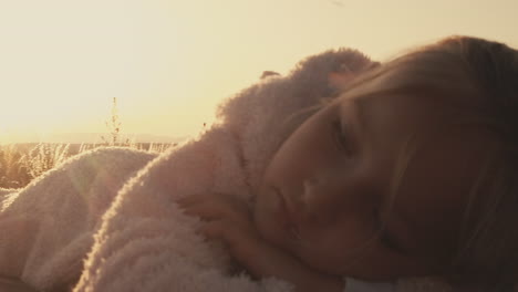 석양이 그녀의 뒤에서 빛나면서 피크닉 테이블에 따뜻한 퍼지 재킷에 껴안은 어린 소녀 주위를 돌아 다니십시오.