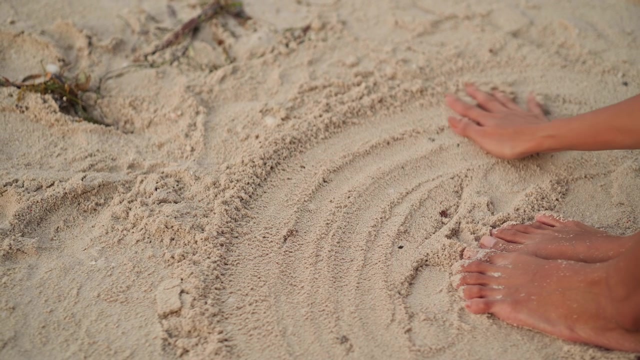 Piedi e mani sulla sabbia della spiaggia che creano un motivo circolare a mandala con lenti movimenti leviganti, granuli testurizzati e piccole alghe, primo piano intimo di un rituale a piedi nudi, luce calda e soffusa e tranquillità