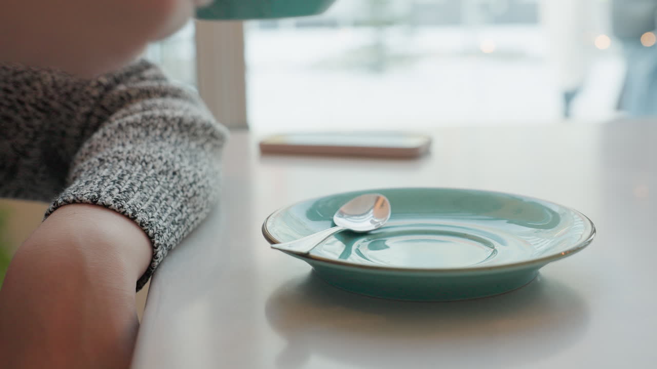 Young fair woman lifting breakfast drink inside cybercafe while people exit through door in winter environment with snow-covered view visible through transparent window, creating peaceful indoor
