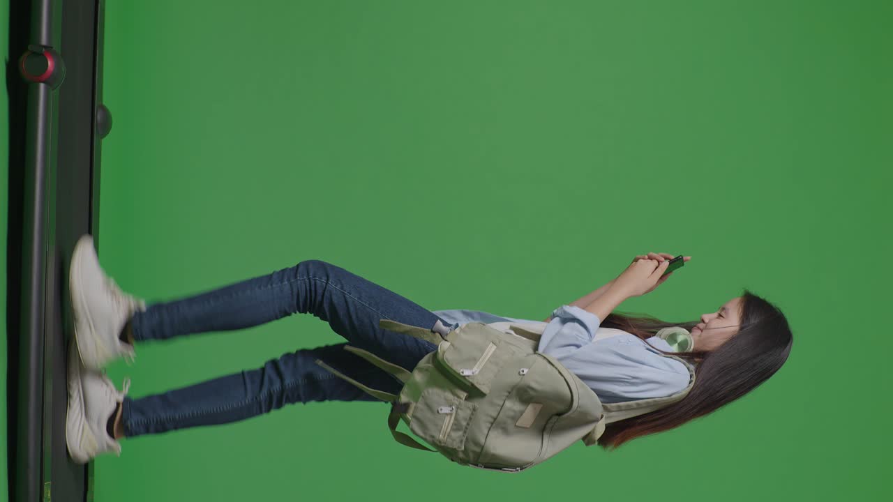 cuerpo lleno de vista lateral estudiante adolescente asiática con una mochila sonriendo y usando un teléfono inteligente mientras camina en la cinta de correr en el estudio de fondo de pantalla verde