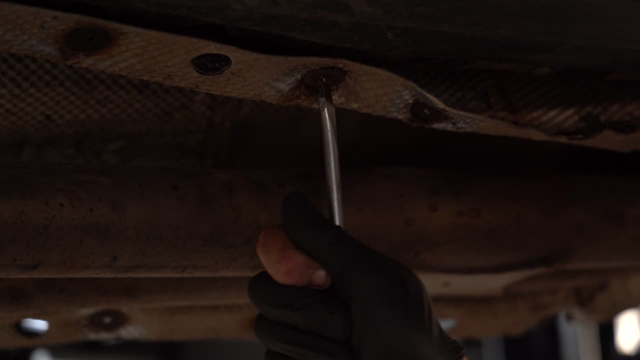A mechanic using a screwdriver to loosen a damaged screw under a vehicle