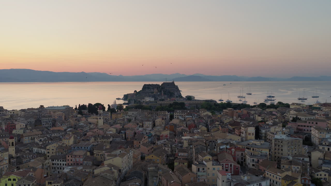 Slow panning drone shot of Corfu old town and the old fortress at dawn, with vibrant pre-sunrise colors and swallows flying in the sky