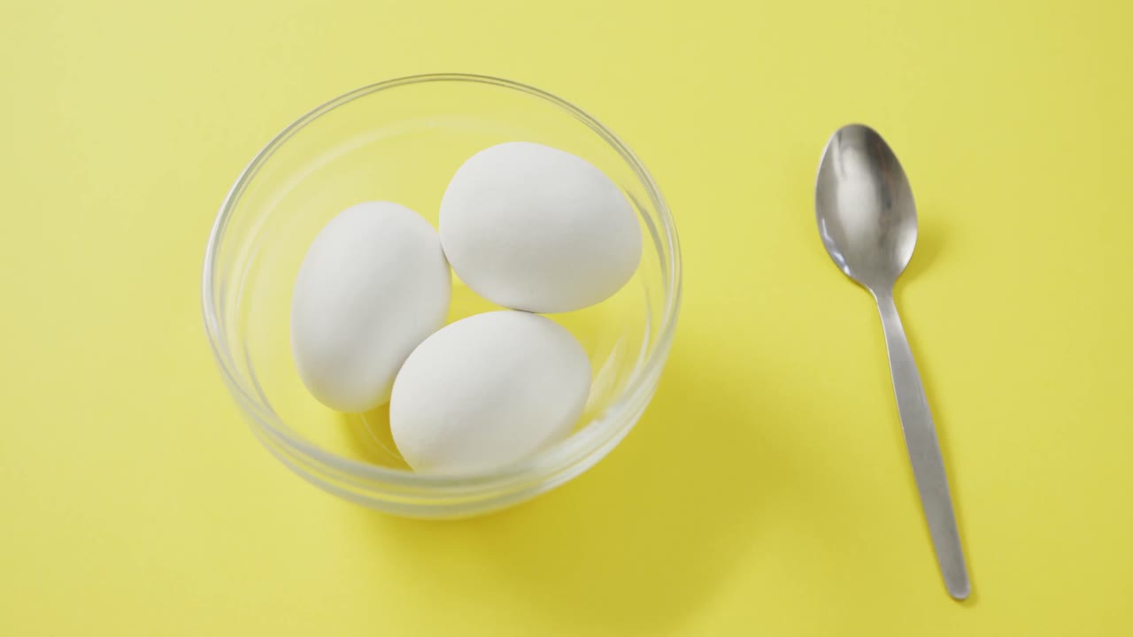 Video of overhead view of glass bowl with eggs and silver spoon on yellow background