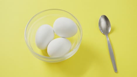 Video-of-overhead-view-of-glass-bowl-with-eggs-and-silver-spoon-on-yellow-background