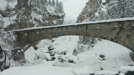 Drohne-Fliegt-Unter-Einer-Steinbrücke-In-Einer-Schneebedeckten-Landschaft